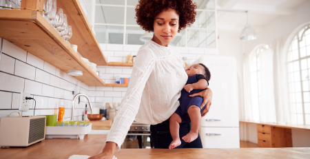 A busy woman holding a baby wipes down a kitchen counter