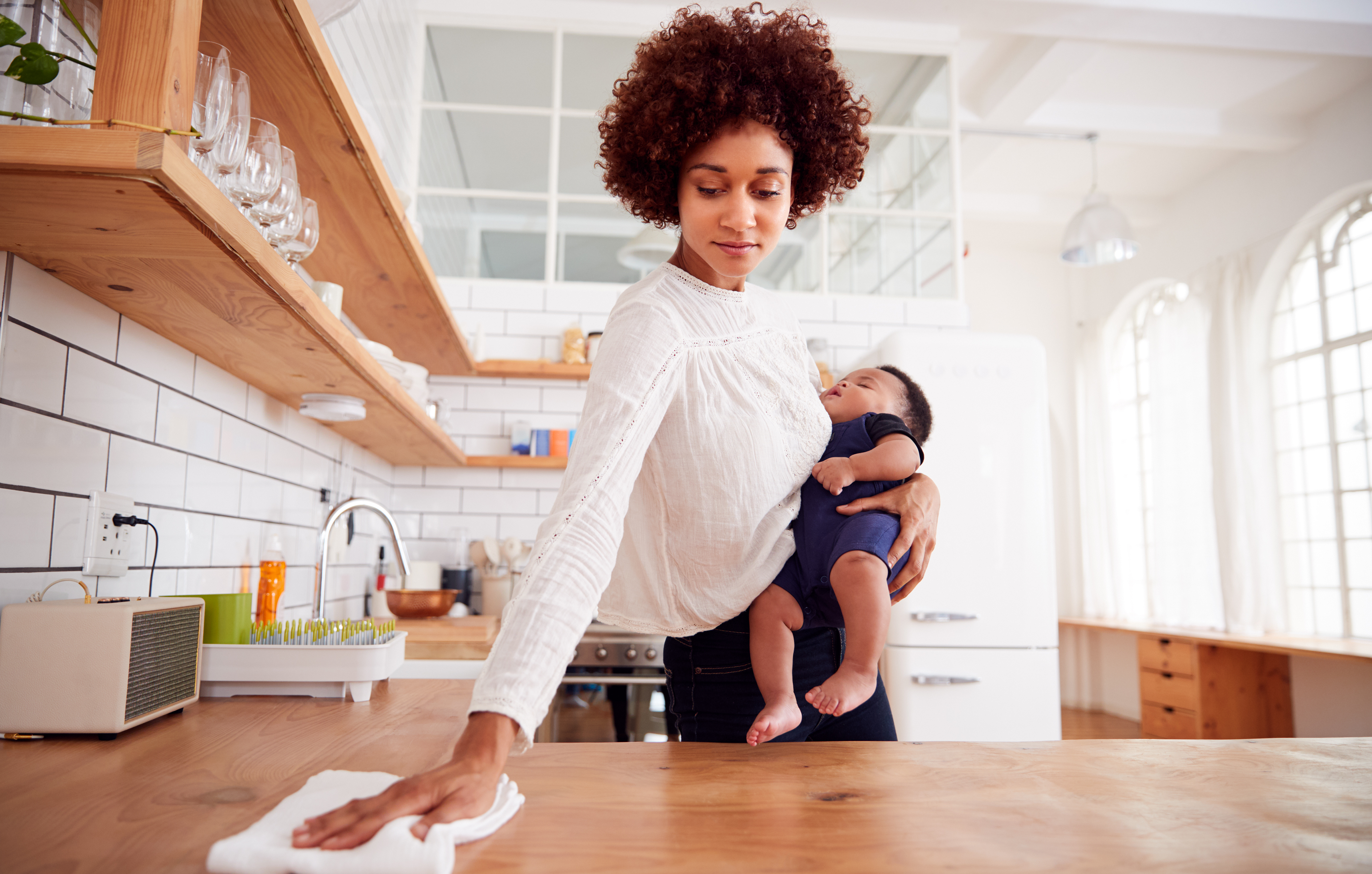 A busy woman holding a baby wipes down a kitchen counter