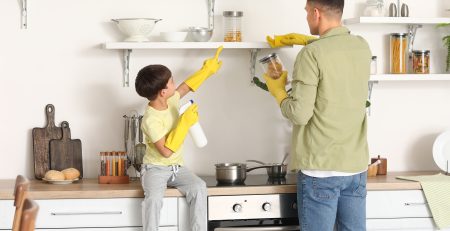 a father and young son wearing rubber gloves and cleaning their kitchen