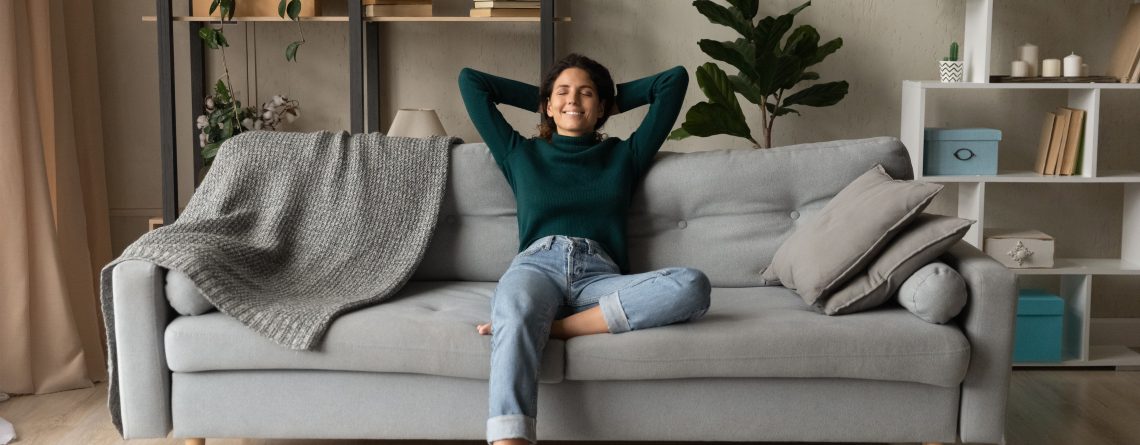 Woman in jeans and green sweater lounging on a grey couch in a clean home. The woman is happy and relaxed.